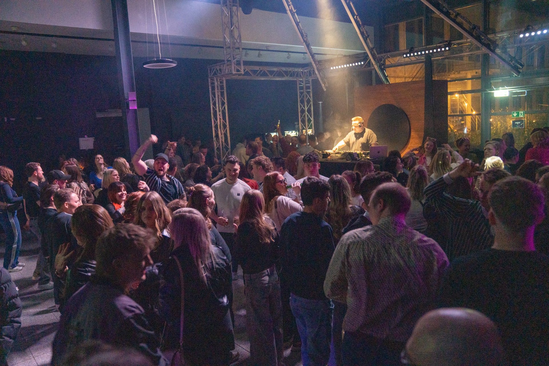 Large crowd of people at an indoor concert or nightclub event with stage lighting and performers visible in the background
